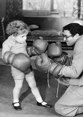Old black and white photo of a small child in big boxing gloves facing a kneeling man in gloves