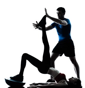 Silhouette of a trainer in a blue shirt holding up the leg of a woman lying on a balance ball, on white